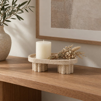 Oval pale beige pedestal tray on two fluted legs on a wooden console holding a cream pillar candle and a small bundle of dried flowers, with a vase of leafy stems and framed art behind