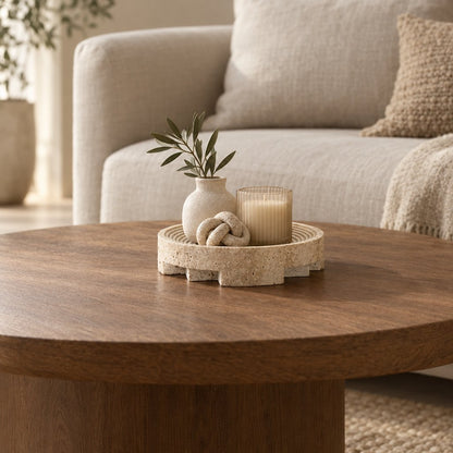Round pale beige pedestal tray on a round wooden coffee table holding a small white vase with an olive branch, a candle in a glass and a knot shaped decor piece, with a cream sofa behind