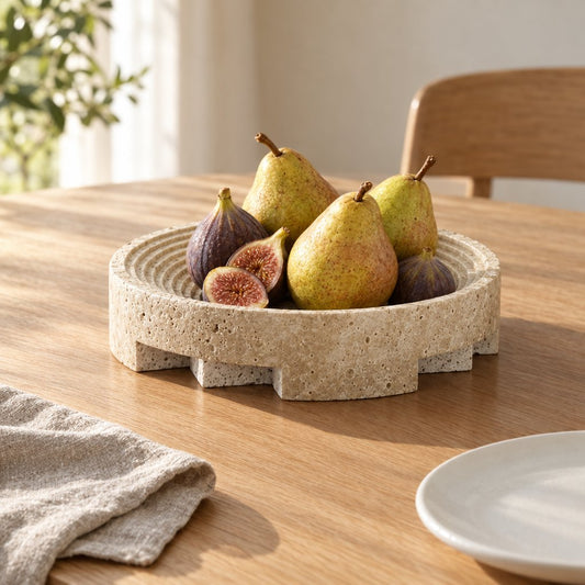 Round pale beige pedestal tray with a ridged inner dish and notched base holding pears and figs, one fig cut open, on a wooden dining table with a gray napkin and white plate