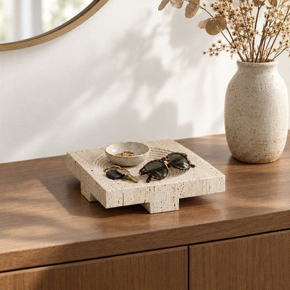 Square pale beige pedestal tray on a wooden console holding a small bowl, sunglasses and keys, beside a speckled vase of dried stems and a round mirror
