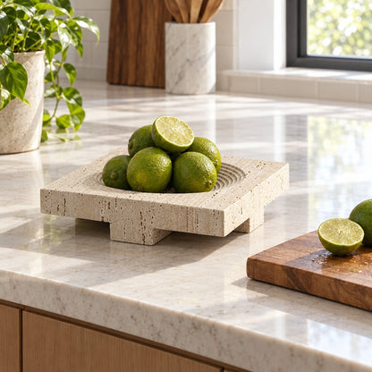 Square pale beige pedestal tray on a light kitchen counter filled with limes, with a potted plant, a halved lime and a wooden board nearby
