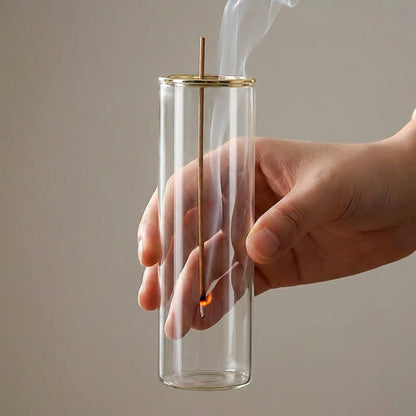Hand holding a clear glass container with a gold rim and a burning incense stick inside, against a neutral background.