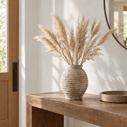 Large textured cream ridged vase with tall pale feathery plumes on a wooden console near a door, with a round mirror above and a wooden tray at right