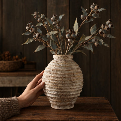 Large textured cream ridged vase with branches of gray green leaves and round seed pods on a dark wood table, with a hand resting on the vase and a dark wood plank wall behind