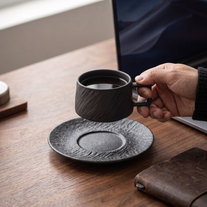 Hand holding a black textured ceramic cup over a matching saucer on a wood desk beside a laptop and a leather notebook