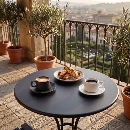 Black and white textured ceramic cups and saucers with coffee and biscotti on a round black table on a balcony with potted olive trees and a view of a garden below