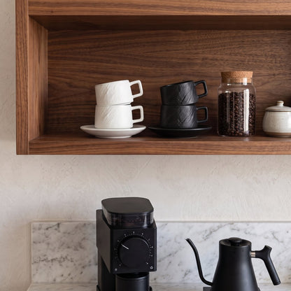 Stacked white and black textured ceramic cups on saucers on a wood shelf above a coffee grinder and a black gooseneck kettle on a marble counter
