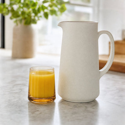 Amber translucent drinking glass of orange juice beside a cream ceramic pitcher on a marble kitchen counter
