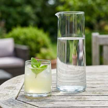 Clear drinking glass of lemonade with ice and a mint sprig beside a glass carafe on a weathered patio table