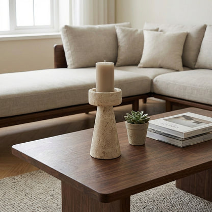 Large travertine pillar candleholder with a taupe pillar candle on a dark wood coffee table beside a small succulent and stacked books with a sofa behind