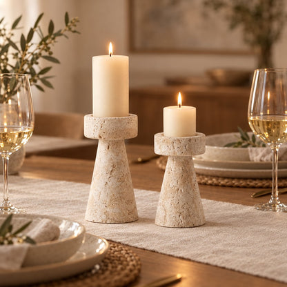 Large and medium travertine pillar candleholders with lit candles on a linen table runner between wine glasses, plates and olive branches on a dining table