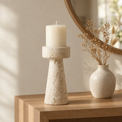 Large travertine pillar candleholder with a cream pillar candle on a wooden console beside a speckled vase with dried stems and a round mirror