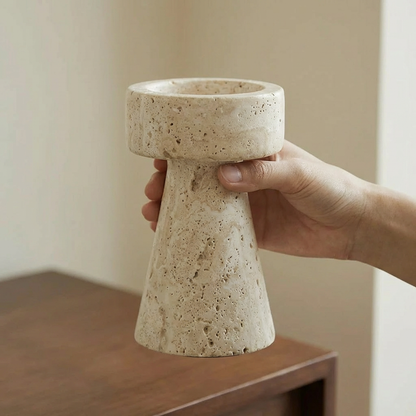 Hand holding a cream travertine pillar candleholder with no candle above a dark wood table
