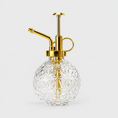 Clear cut glass round plant mister with a gold pump top, spout and ring handle on a light grey background