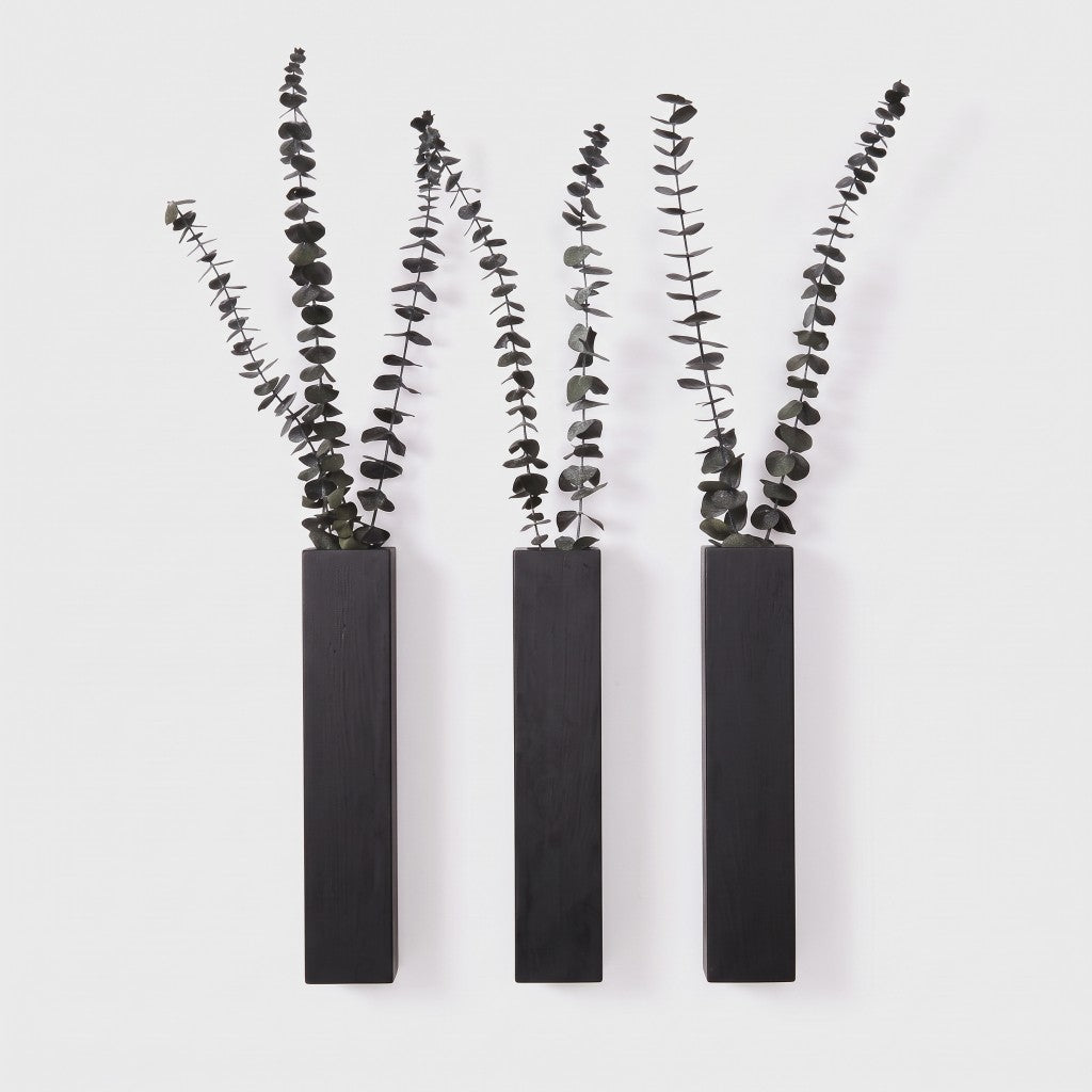 Three narrow black wall planters holding dark leafy eucalyptus stems on a plain light background