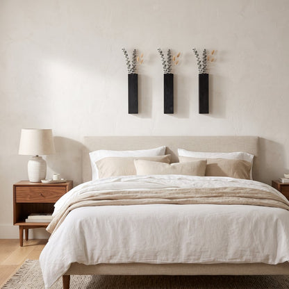 Three narrow black wall planters holding dried stems on a cream wall above a bed with white bedding and a nightstand lamp