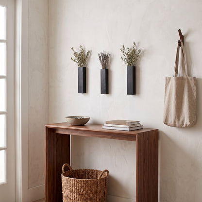 Three narrow black wall planters with dried stems on a cream wall above a wood console table with a bowl, books and a basket, with a canvas tote hanging on a hook