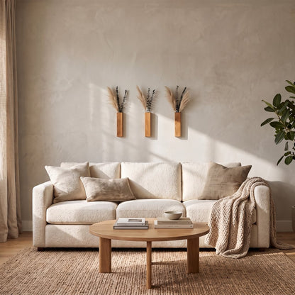 Three narrow brown wood wall planters with dried stems on a cream wall above a cream sofa with pillows, a throw and a round wood coffee table