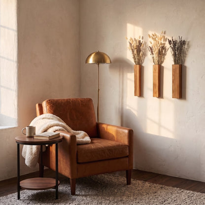 Three narrow brown wood wall planters with dried stems on a cream wall beside a brass floor lamp, a tan leather armchair with a throw and a small side table
