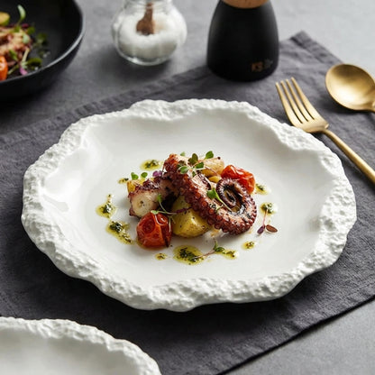 White ceramic plate with a textured rim holding grilled octopus with potatoes and cherry tomatoes on a dark grey linen mat, with gold cutlery and a pepper mill