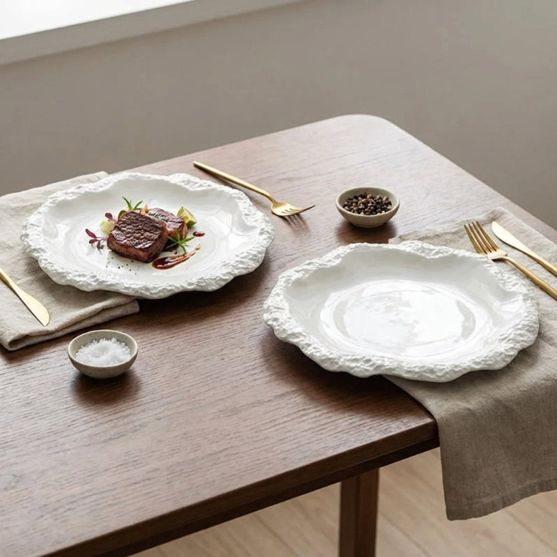 White ceramic plates with decorative edges on a wooden table with gold cutlery and small bowls.