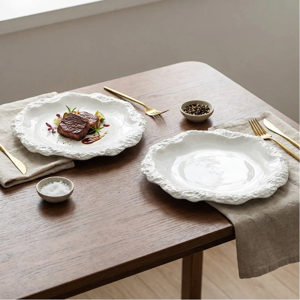 Two white ceramic plates with a textured rim holding a small steak dish on a wood table, with gold cutlery, small salt and pepper dishes and a beige napkin
