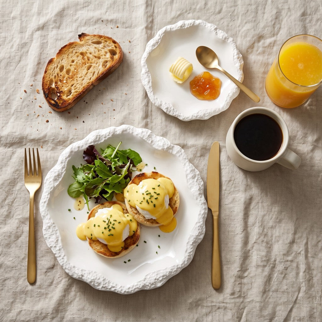 Overhead view of a white ceramic plate with a textured rim holding eggs Benedict with greens, beside a smaller plate with butter and jam, toast, orange juice, black coffee and gold cutlery on a linen cloth