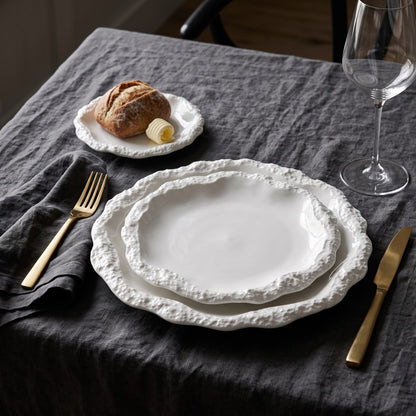 Two stacked white ceramic plates with a textured rim on a dark grey linen cloth, with a small side plate holding bread, gold cutlery and a wine glass