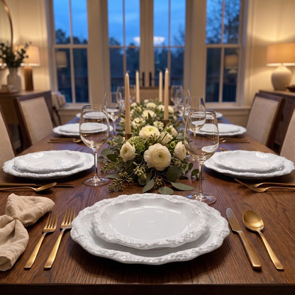 Place setting with stacked white ceramic plates with a textured rim, gold cutlery and glasses on a dark wood table with a white rose centerpiece and tall candles