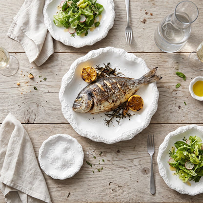 Overhead view of a white ceramic plate with a textured rim holding a whole grilled fish with lemon on a weathered wood table, with salad plates, a small dish, a glass pitcher and linen napkins
