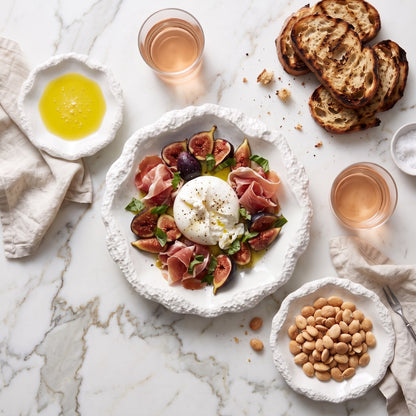 Overhead view of white ceramic plates with a textured rim holding burrata with prosciutto and figs, a small dish of olive oil and a bowl of nuts, with grilled bread and glasses on a white marble surface