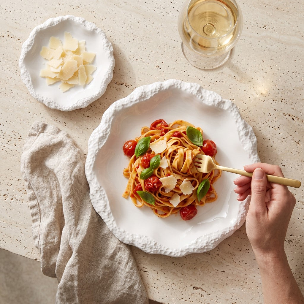 Hand twirling pasta with cherry tomatoes and basil on a white ceramic plate with a textured rim, beside a small plate of shaved parmesan and a glass of white wine on a stone table
