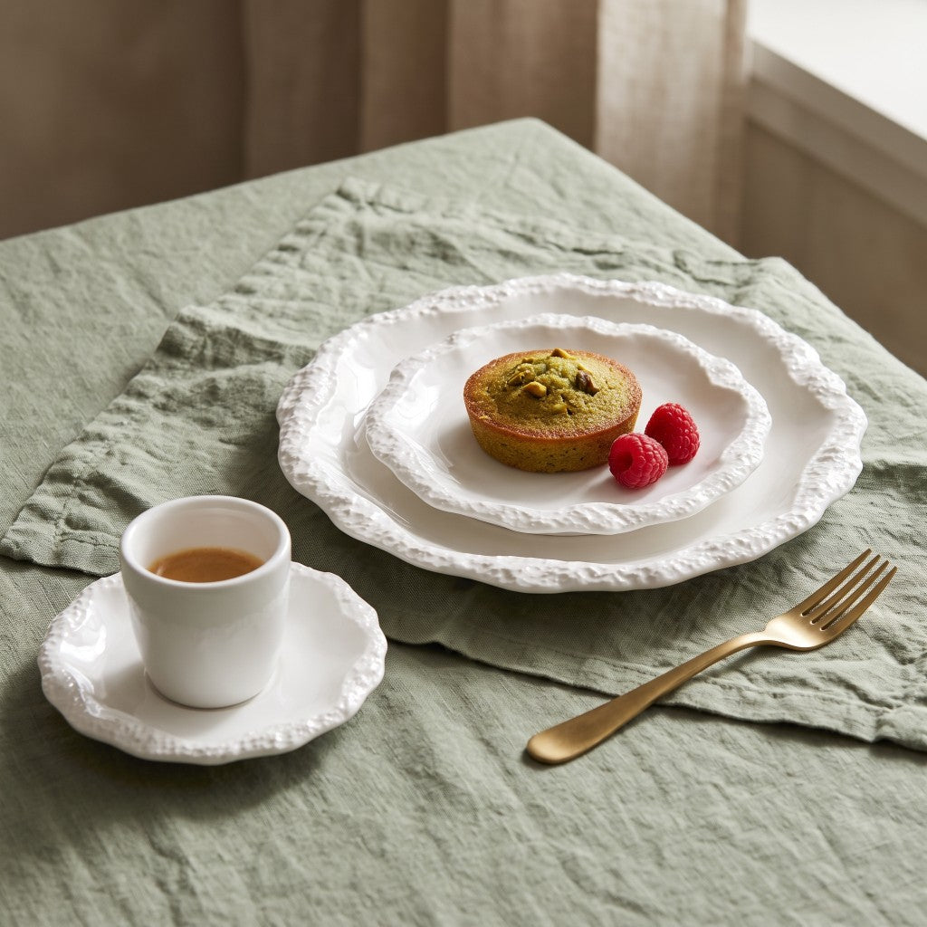 Two stacked white ceramic plates with a textured rim holding a small pistachio-topped cake with two raspberries, beside a white cup and saucer and a gold fork on a sage green linen cloth