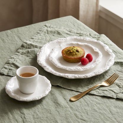 Two stacked white ceramic plates with a textured rim holding a small pistachio-topped cake with two raspberries, beside a white cup and saucer and a gold fork on a sage green linen cloth