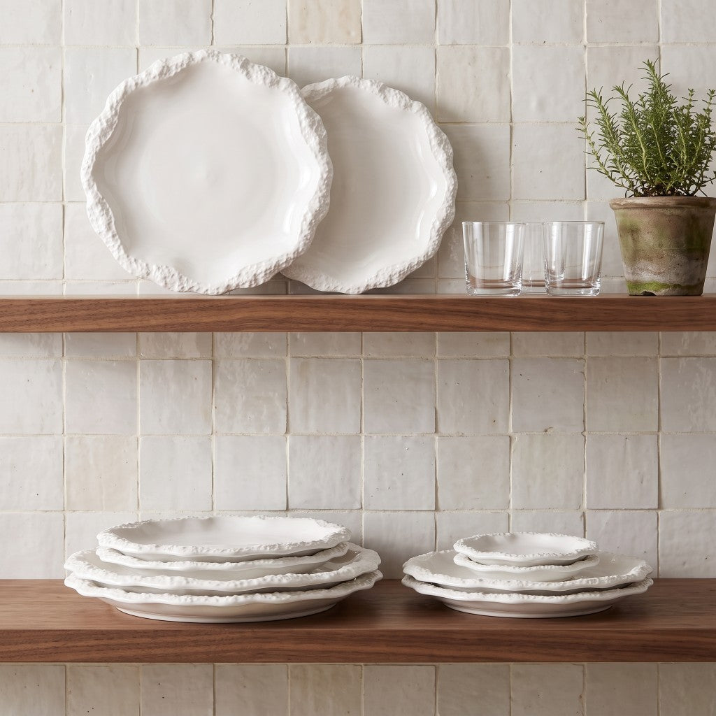 Two white ceramic plates with a textured rim standing on a wood shelf against a cream tile wall, with stacked plates below, glasses and a potted herb