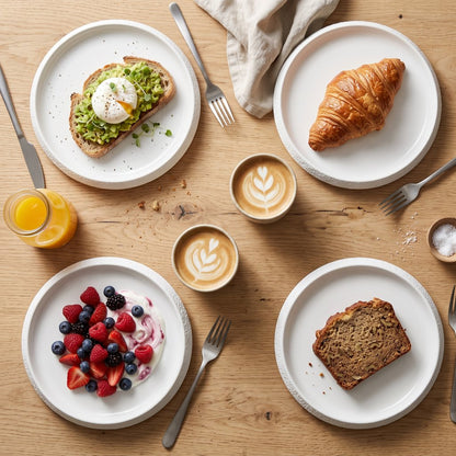 Overhead view of four white textured ceramic plates holding avocado toast with a poached egg, a croissant, berries with yogurt and a slice of bread, with lattes, orange juice and cutlery on a wood table