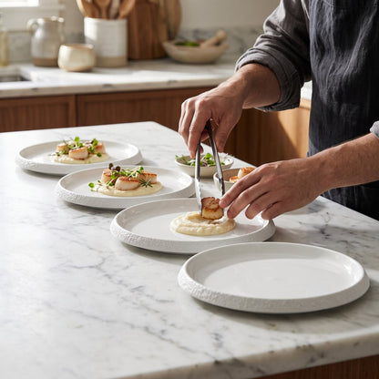 Hands plating scallops with sauce onto white textured ceramic plates lined up on a marble kitchen counter
