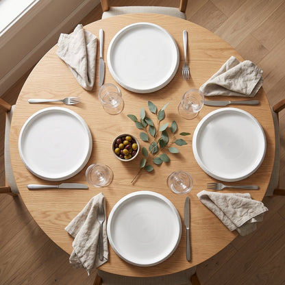 Overhead view of a round wood table set with four white textured ceramic plates, glasses, linen napkins and a small bowl of olives with a leafy sprig