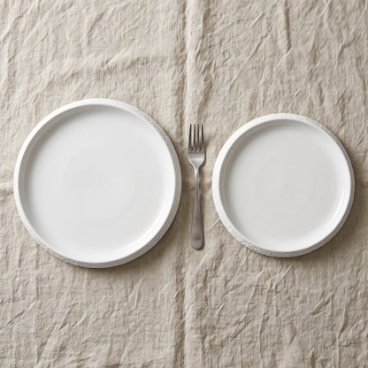 Two white textured ceramic plates in different sizes with a fork between them on a beige linen cloth