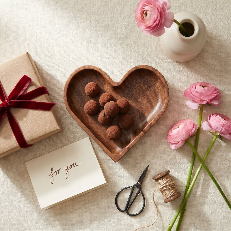 Heart-shaped wooden dish with chocolate truffles, a gift box, pink flowers, and a card on a light surface.