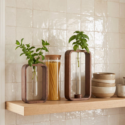 Two dark brown wood frame vases in different heights with glass tubes holding leafy green sprigs on a light wood shelf against a cream tile wall, beside a glass jar with a wood lid and stacked ceramic bowls
