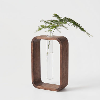 Dark brown wood frame vase in a rounded rectangle shape with a glass tube holding a feathery green sprig, on a light grey background