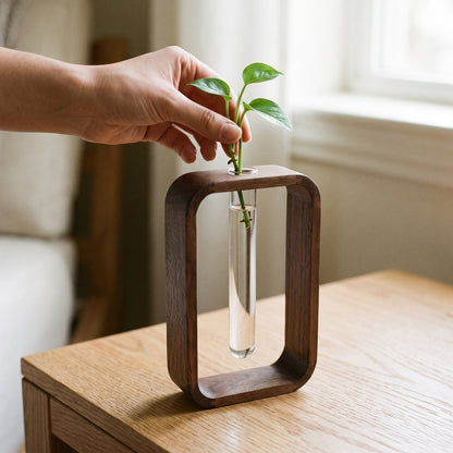 Hand placing a small green plant cutting into the glass tube of a dark brown wood frame vase on a light wood table by a window