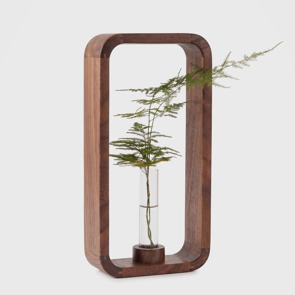 Tall dark brown wood frame vase in a rounded rectangle shape with a glass tube on a round wood base holding a feathery green sprig, on a light grey background
