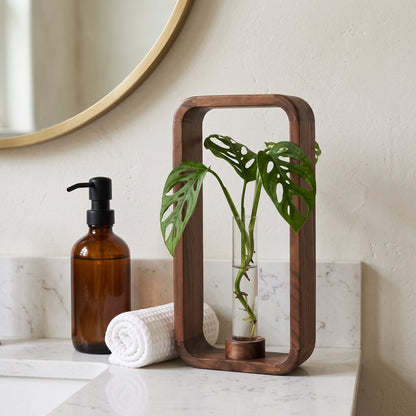 Tall dark brown wood frame vase with a glass tube holding a green plant cutting with split leaves on a white marble-look bathroom vanity beside an amber glass soap pump bottle and a rolled white towel
