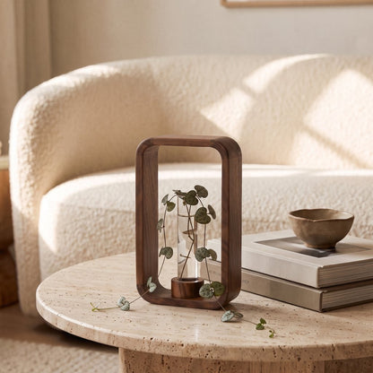 Dark brown wood frame vase with a glass tube holding a trailing vine with small round green leaves on a round pale stone-look coffee table with stacked books and a small bowl, in front of a cream boucle sofa