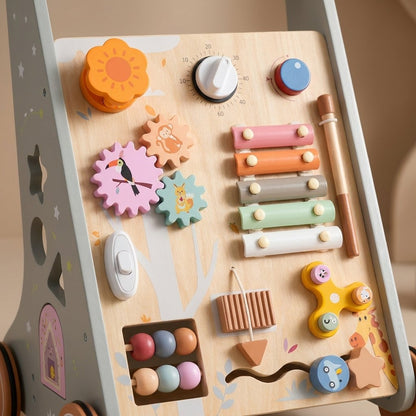 A wooden sensory and motor play table with colorful interactive toys, including gears, a xylophone, fidget spinner, beads, and a moon face button, shown in a warm beige room setting.