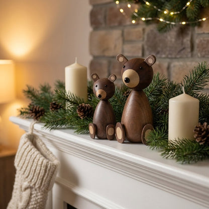 Small and large wooden bear figurines on a white mantel with a pine garland, pinecones, and cream candles