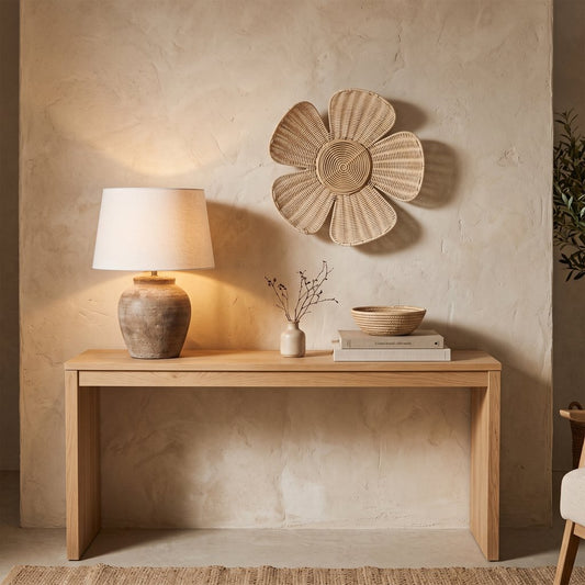 Woven Bloom flower wall decor in light tan on a beige plaster wall above a light wood console table with a lamp, a small vase with dried stems, and books with a bowl