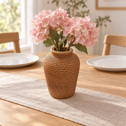 Woven rattan vase with a flared rim holding pink hydrangeas on a linen runner on a wooden dining table between white plates